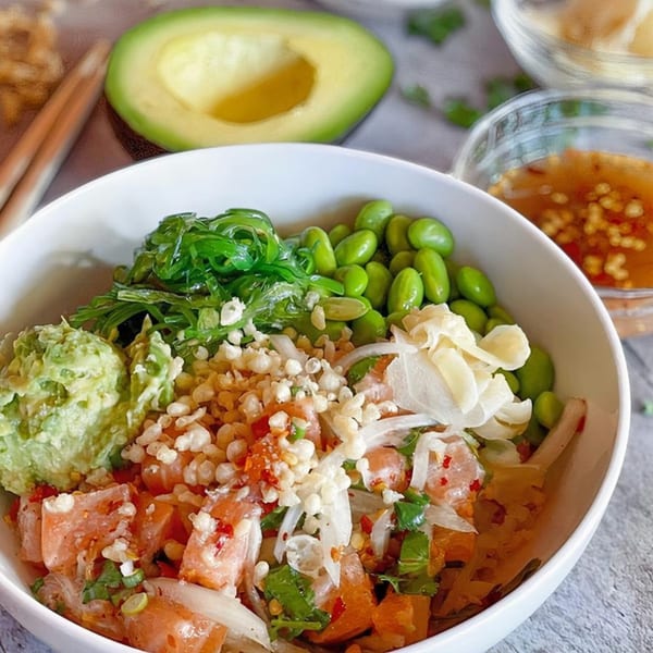 A vibrant poke bowl with salmon, avocado, edamame, seaweed, and crispy toppings, with an avocado half.