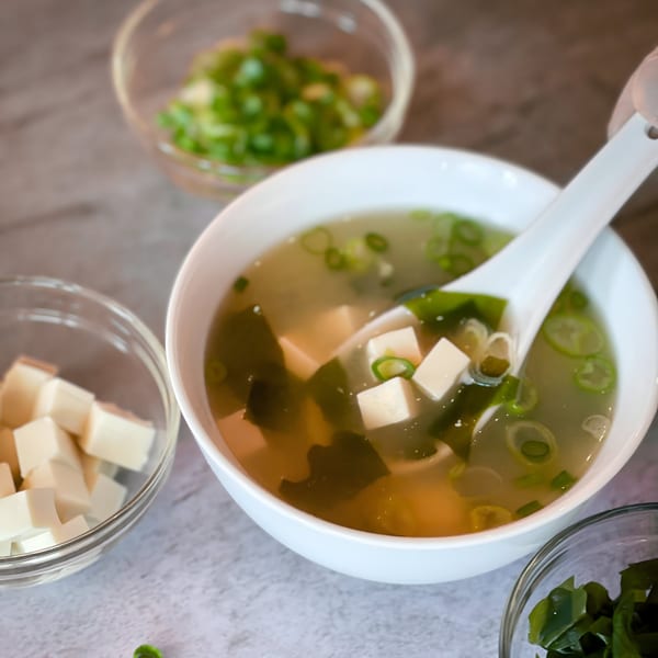 A white bowl of miso soup with tofu, seaweed, and green onions, with ingredients in separate bowls.